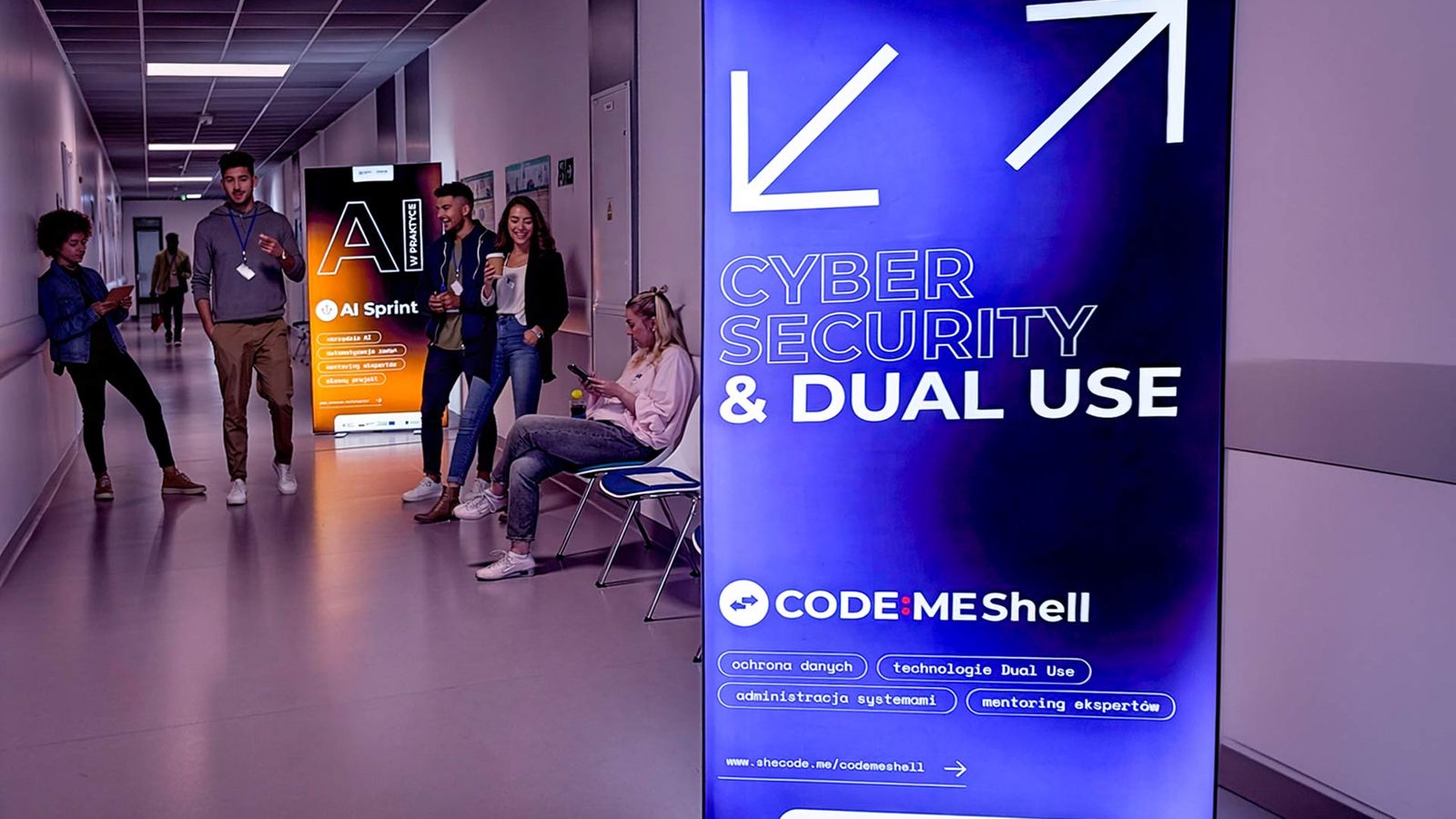 An illuminated “Cybersecurity & Dual Use” banner of the CODE:ME Shell program, with participants in the faculty corridor behind it.